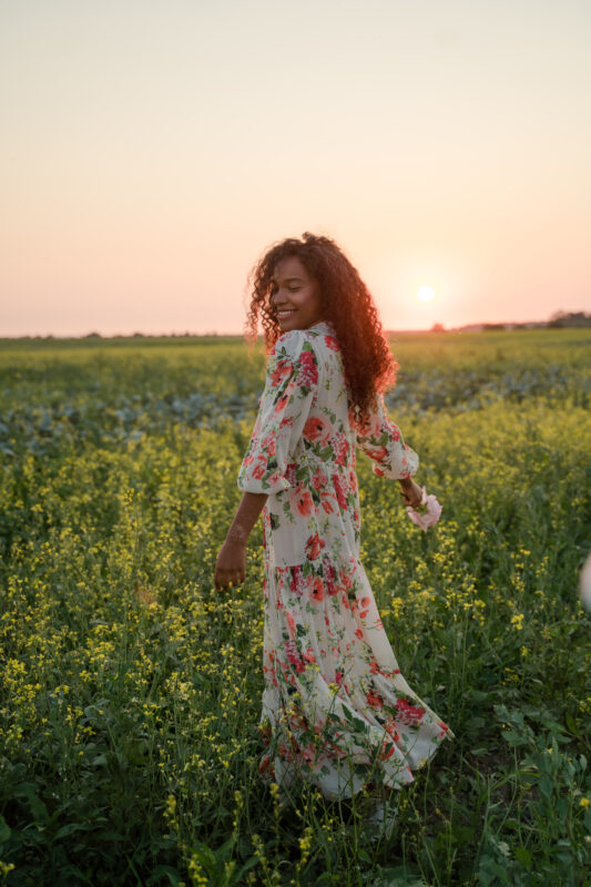 Cheerful woman in floral summer dress dancing in flowery meadow at sunset